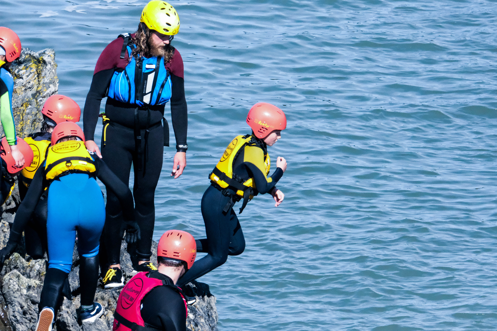 Zdjęcie przedstawia grupę ludzi w różnym wieku,  którzy ubrani są w pianki do pływania, kapoki oraz kaski ochronne. Wszyscy stoją na skalistym brzegu tuż nad taflą wody. Jedna z osób przygotowuje się do skoku i przechyla się w kierunku wody. 