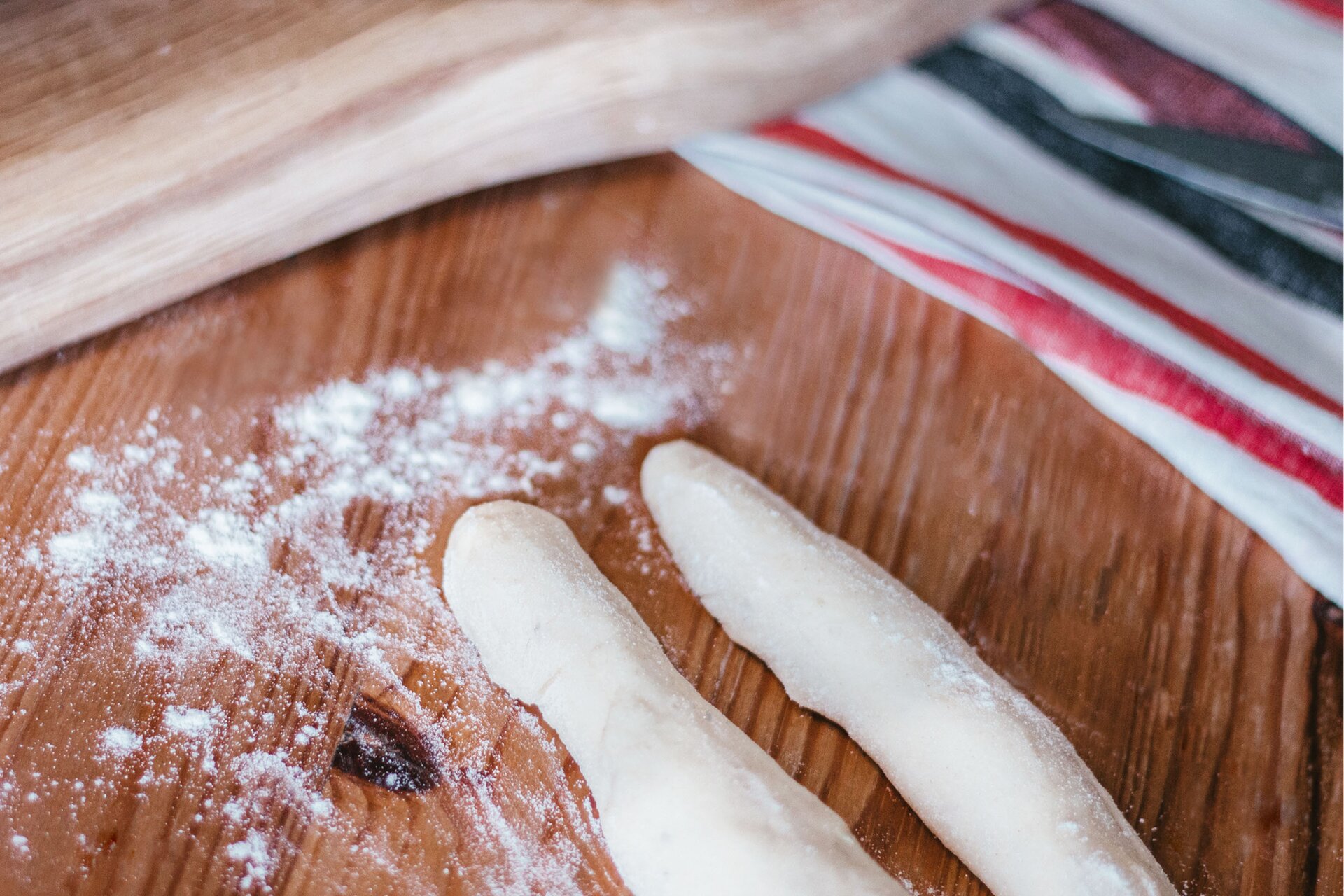 Zdjęcie przedstawia krok drugi formowania rogali węgierskich. Ujęcie wykonane jest od góry pod skosem. Na pierwszym planie w dolnej środkowej części grafiki znajdują się dwa, równej długości wałki ciasta oprószone białą mąką. Leżą one na ciemnobrązowym drewnianym blacie. Dookoła nich rozsypana jest mąka. Na drugim planie w prawym rogu zdjęcia dostrzec można fragment białej ścierki z pasami w kolorze czerwonym i szarym. W lewym górnym rogu grafiki widoczna jest drewniana jasnobrązowa deska leżąca częściowo na blacie, a częściowo na ścierce.