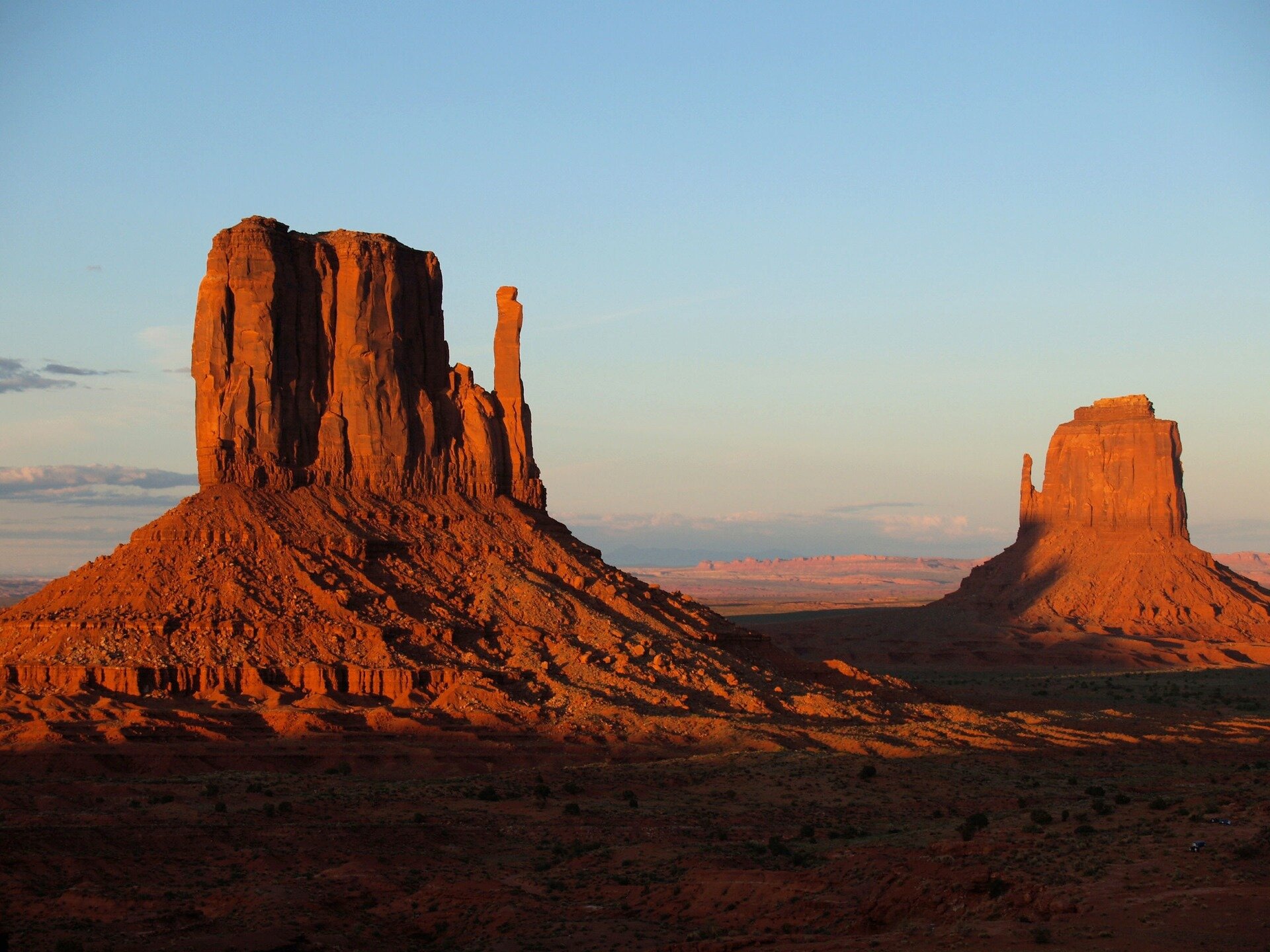 Fotografia prezentuje pustynię skalistą w Monument Valley w USA. Na pierwszym planie widoczne dwie wysokie, prostokątne góry u podstawy, których znajdują się zwietrzałe okruchy skalne. Skały są koloru czerwonego.