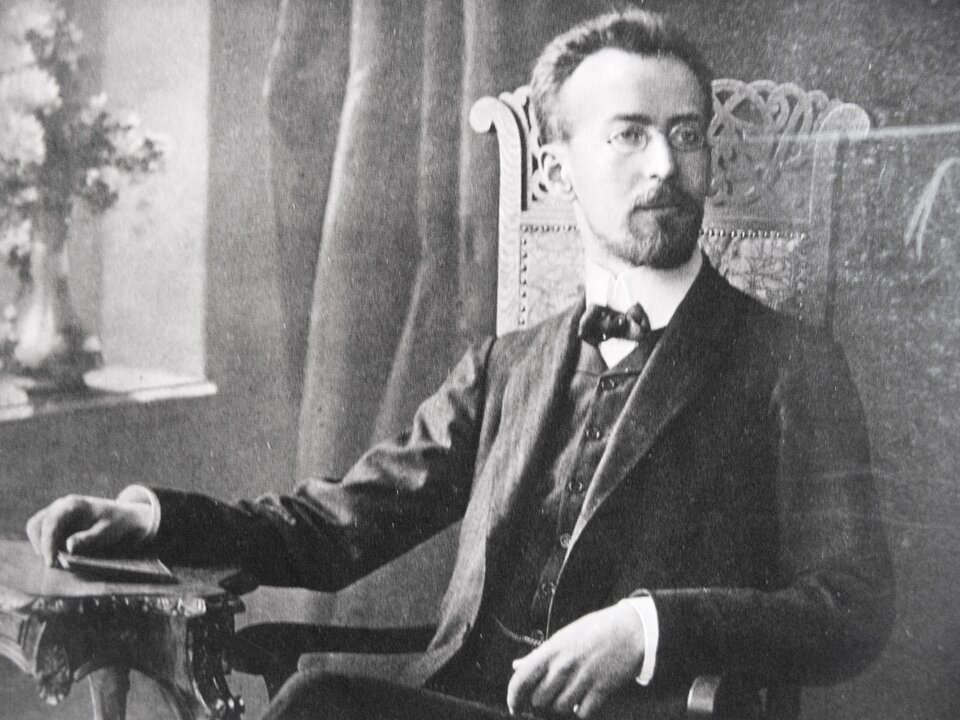 Ilustracja przedstawia polskiego kompozytora i dyrygenta Mieczysława Karłowicza. Mężczyzna na fotografii siedzi na fotelu. Ubrany jest w białą koszulę, z muszką pod szyją, kamizelkę oraz garnitur. Kompozytor ma okulary.