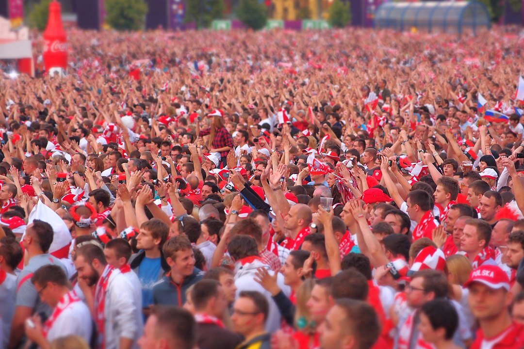 Zdjęcie przedstawia stojący tłum kibiców. Większość z nich ma na szyjach biało‑czerwone szaliki. Część osób ma podniesione do góry ręce.
