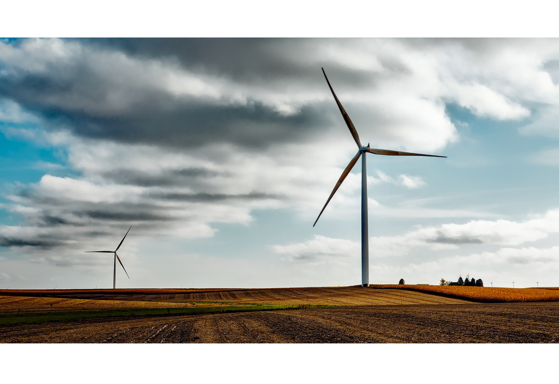Zdjęcie przedstawia dwa wysokie wiatraki, które produkują prąd. Stoją na polu, widoczne są na tle pochmurnego nieba.