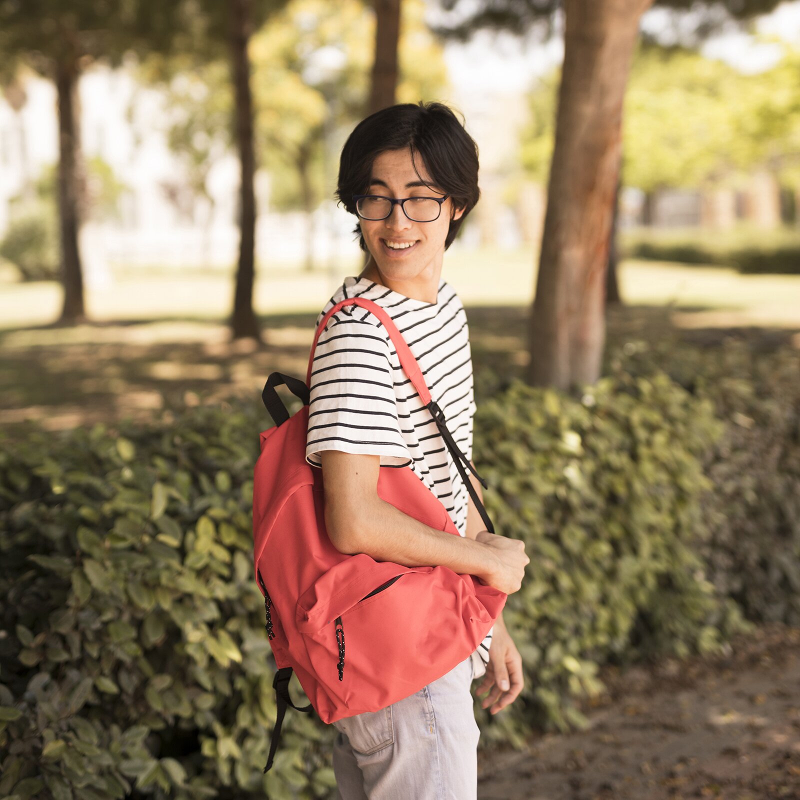 Zdjęcie przedstawia chłopaka, w ujęciu od kolan w górę, stojącego na alejce przy żywopłocie w parku. Chłopak ma ciemne włosy, okulary korekcyjne, jest ubrany w koszulkę z krótkim rękawem w białe i czarne paski oraz szare spodnie. Na prawym ramieniu ma przewieszony czerwony plecak. Chłopak ogląda się za siebie i lekko uśmiecha.
