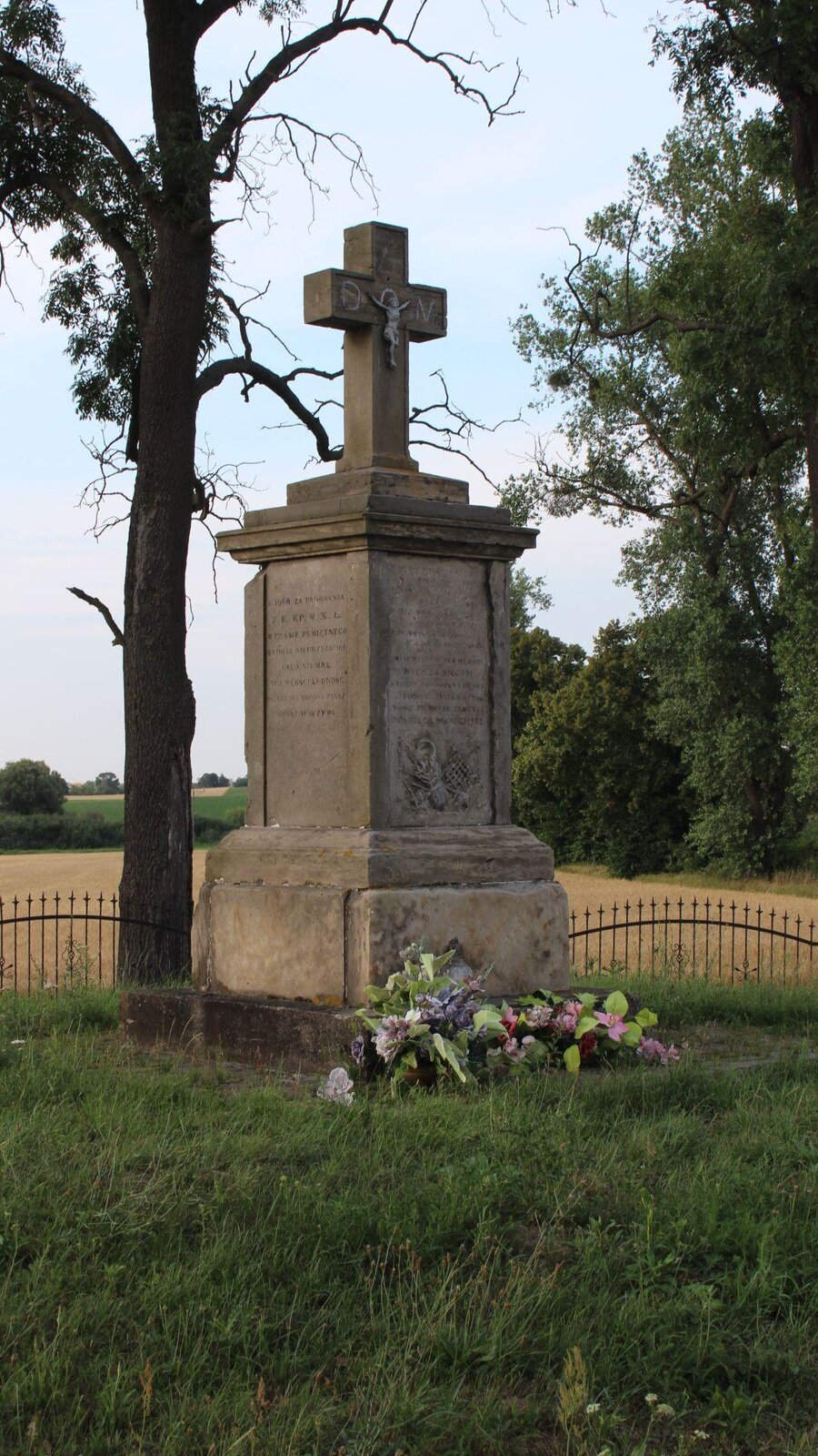 Zdjęcie przedstawia nagrobek przypominający obelisk. Na jego bokach znajdują się wyryte napisy, które są nieczytelne. U szczytu grobu umieszczony został krzyż. Pod pomnikiem ułożone zostały różowe i fioletowe kwiaty. W tle widoczny jest metalowy płot, za którym rosną drzewa, a także pola uprawne. Niebo jest błękitne i bezchmurne. Zdjęcie zostało wykonane w świetle dziennym.