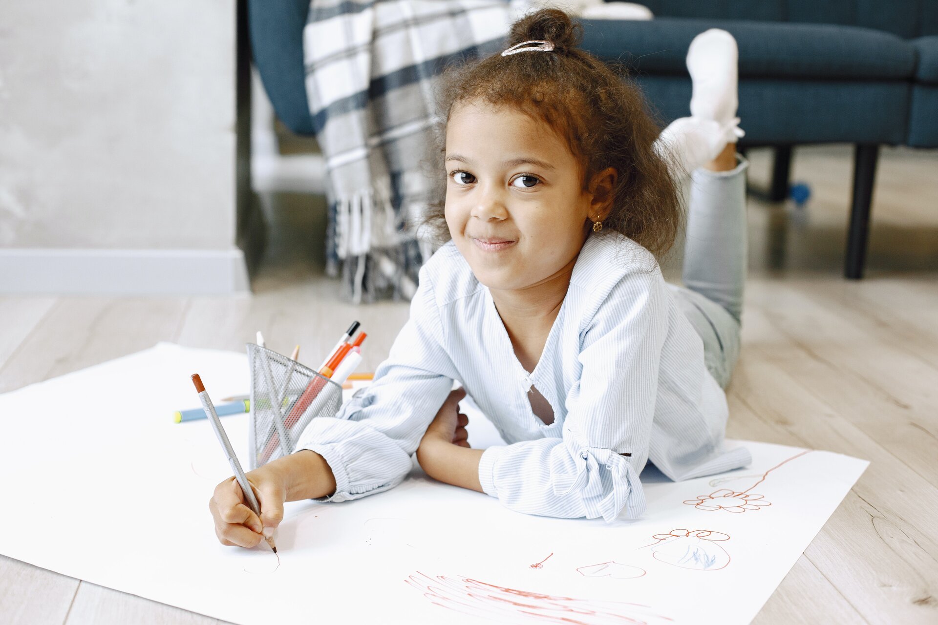 Na ilustracji znajduje się dziewczynka leżąca na podłodze i rysująca kredkami.