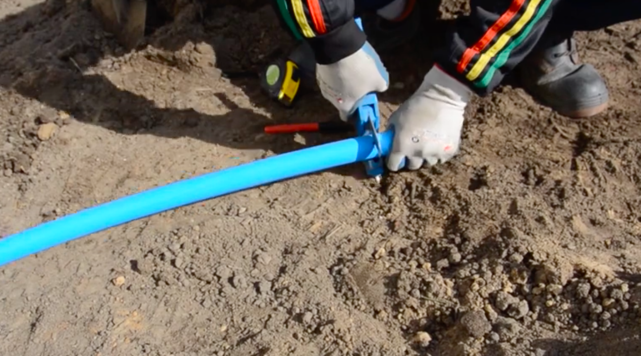 Na zdjęciu widoczne jest cięcie rury z tworzywa sztucznego. Pracownik przecina ją pod kątem prostym trzymanymi w dłoni niewielkimi nożycami. Poniżej ilustracji znajduje się przycisk do odtworzenia nagrania.