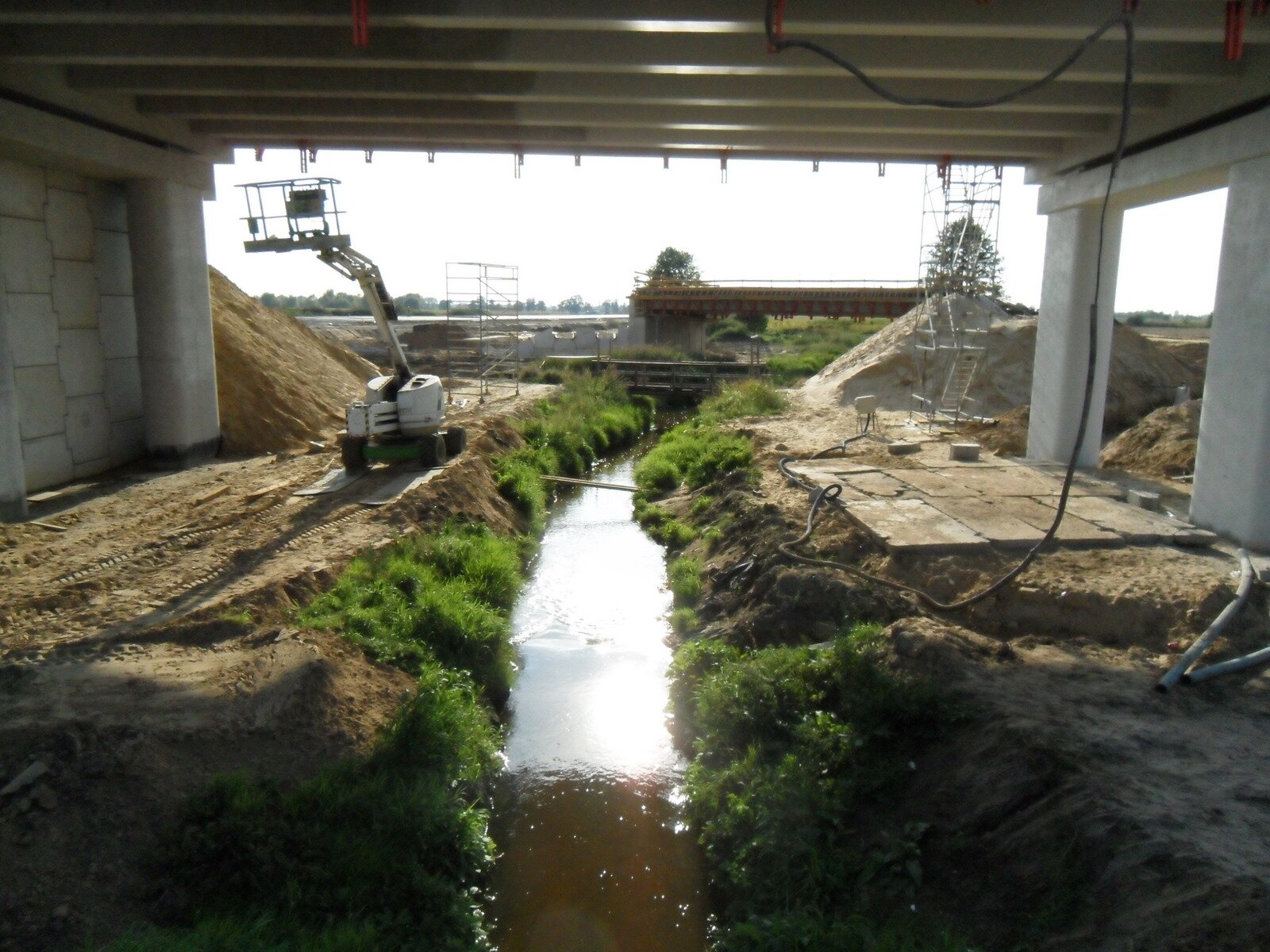 Budowa A2 nad Zimną Wodą na granicy Brwinowa i Biskupic. Zdjęcie przedstawia prace budowlane. Po środku jest rów, w którym znajduje się woda. Obok stoi sprzęt budowlany. 