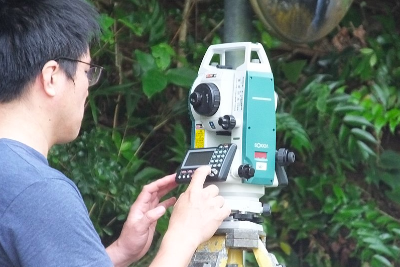 Na zdjęciu widoczny jest mężczyzna obsługujący teodolit. Teodolit ustawiony jest na trójnożnym statywie. Wyposażony jest w lunetę i niewielki prostokątny ekran z wyświetlaczem oraz wieloma przyciskami.