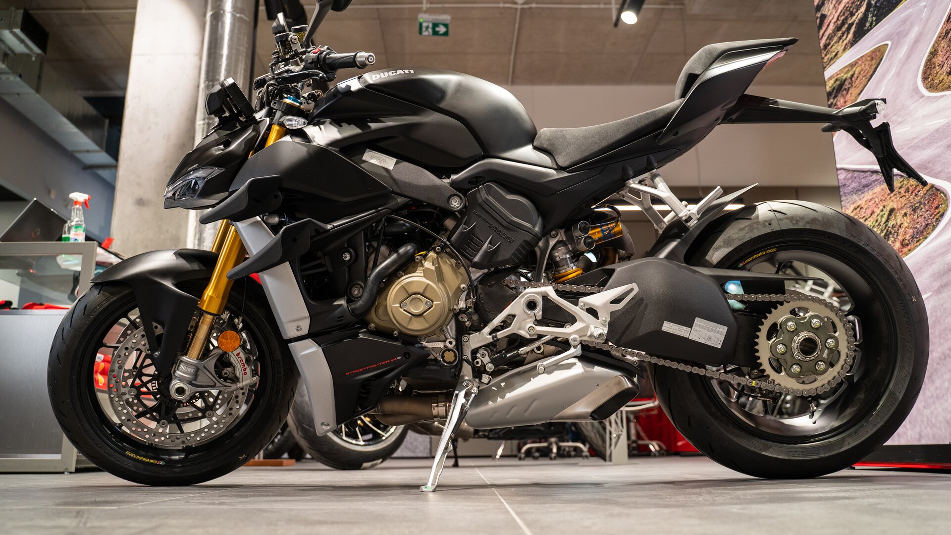 Zdjęcie przedstawia motocykl. Ma sporo odsłoniętych elementów. Jest czarny. Ma złote elementy poniżej kierownicy. Tylny błotnik jest znacznie uniesiony powyżej koła.   