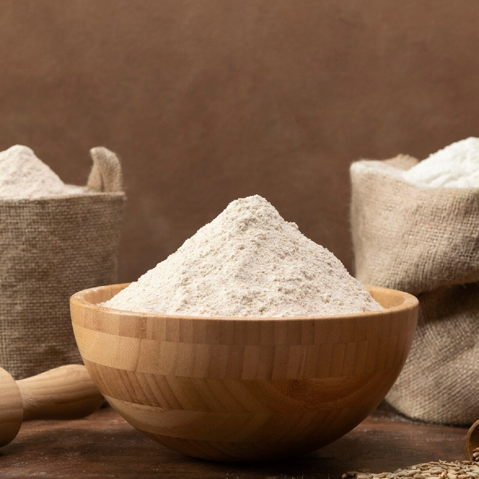 Zdjęcie przedstawia drewnianą miseczkę z mąką. W tle dwa worki z mąką i wałek.