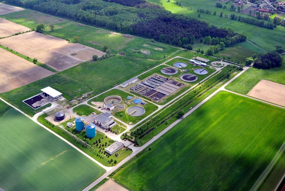 Zdjęcie wykonane z góry na prostokątne pola, pomiędzy którymi jest oczyszczalnia ścieków. W oczyszczalni znajduje się kilka okrągłych oraz prostokątnych zbiorników, dwa silosy oraz budynki. W oddali rośnie las oraz jest zabudowa jednorodzinna.    
