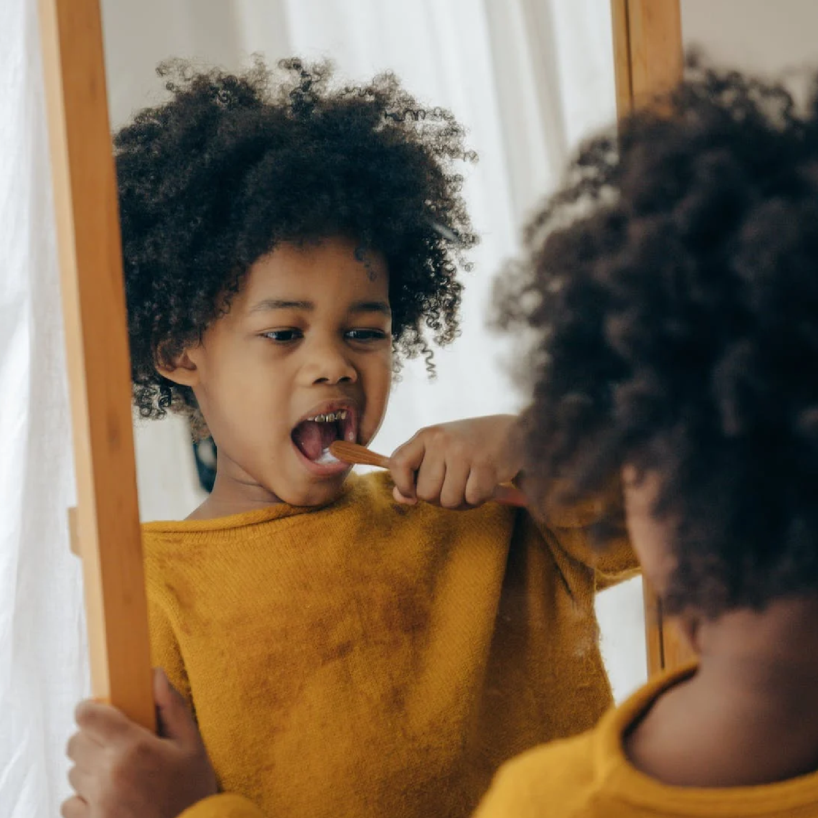 Zdjęcie przedstawia czarnoskórego chłopca w fryzurze afro, który szczotkuje swoje zęby patrząc się w lustro.