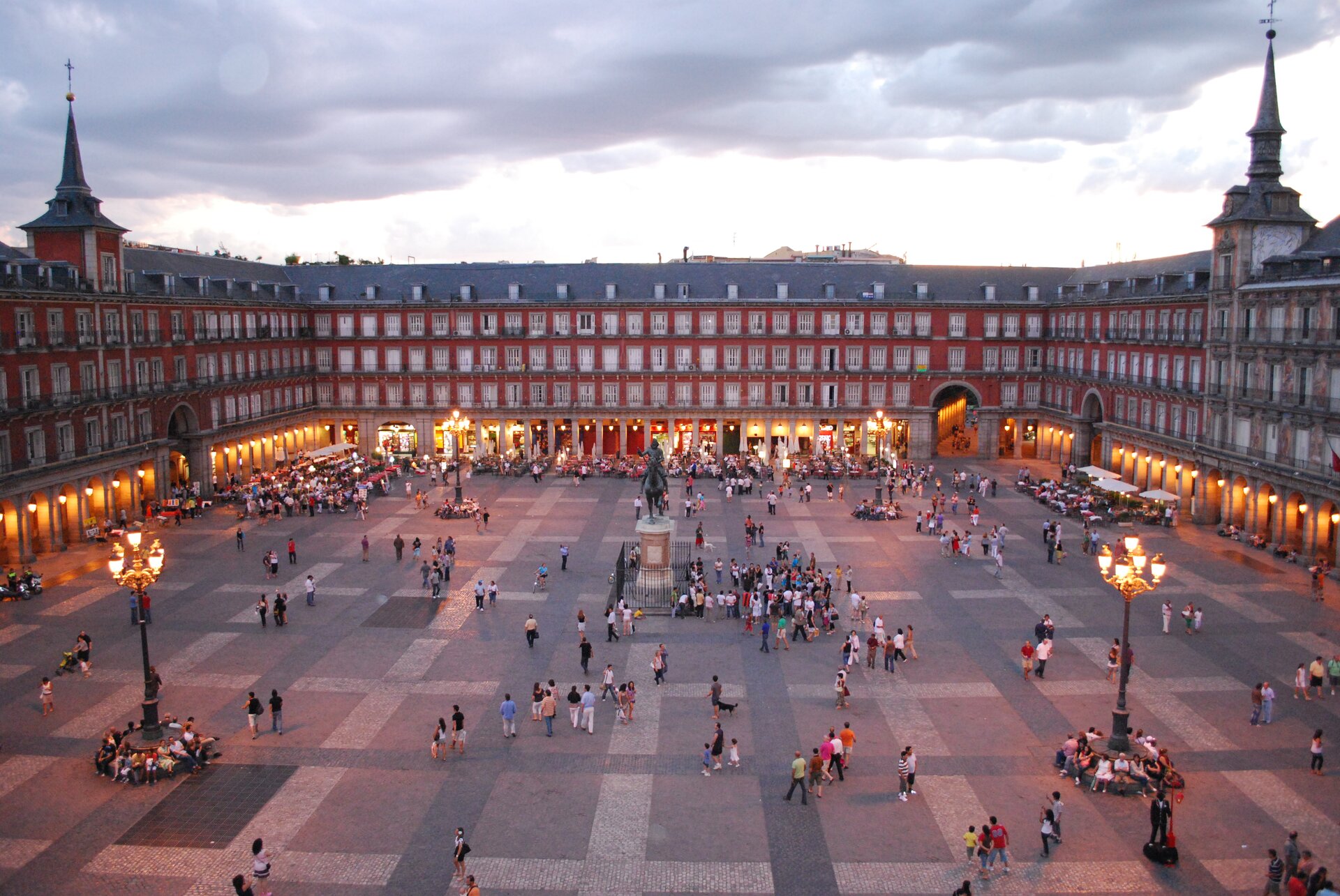 Na zdjęciu prostokątny plac Plaza Mayor w Madrycie otoczony  trzypiętrowymi pomarańczowymi budynkami z dużą ilością balkonów. W kolumnadach palą się światła. W centrum – pomnik przedstawiający jeźdźca na koniu. Na placu stoją cztery symetrycznie ustawione latarnie. Po placu chodzą ludzie. W tle wczesnowieczorne, pogodne niebo.