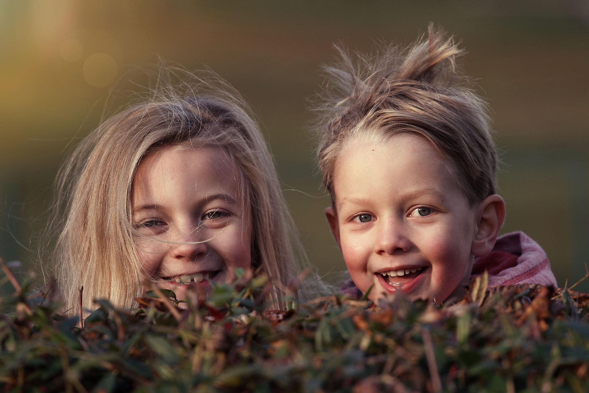 Fotografia przedstawia dwójkę uśmiechających się dzieci , dziewczynkę i chłopca. Dzieci wychylają głowy nad krzewem, mają blond, rozwiane przez wiatr włosy. 