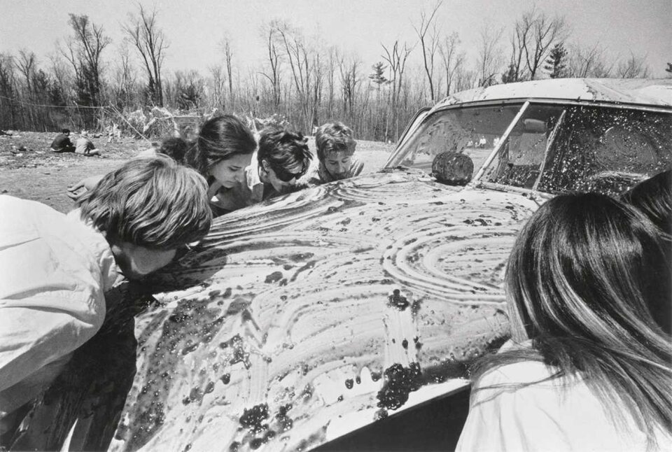 Stare czarno białe zdjęcie przedstawia auto, wokół którego zebrało się kilka kobiet. Maska pojazdu pokryta jest dżemem, który zlizywany jest przez zgromadzone osoby.