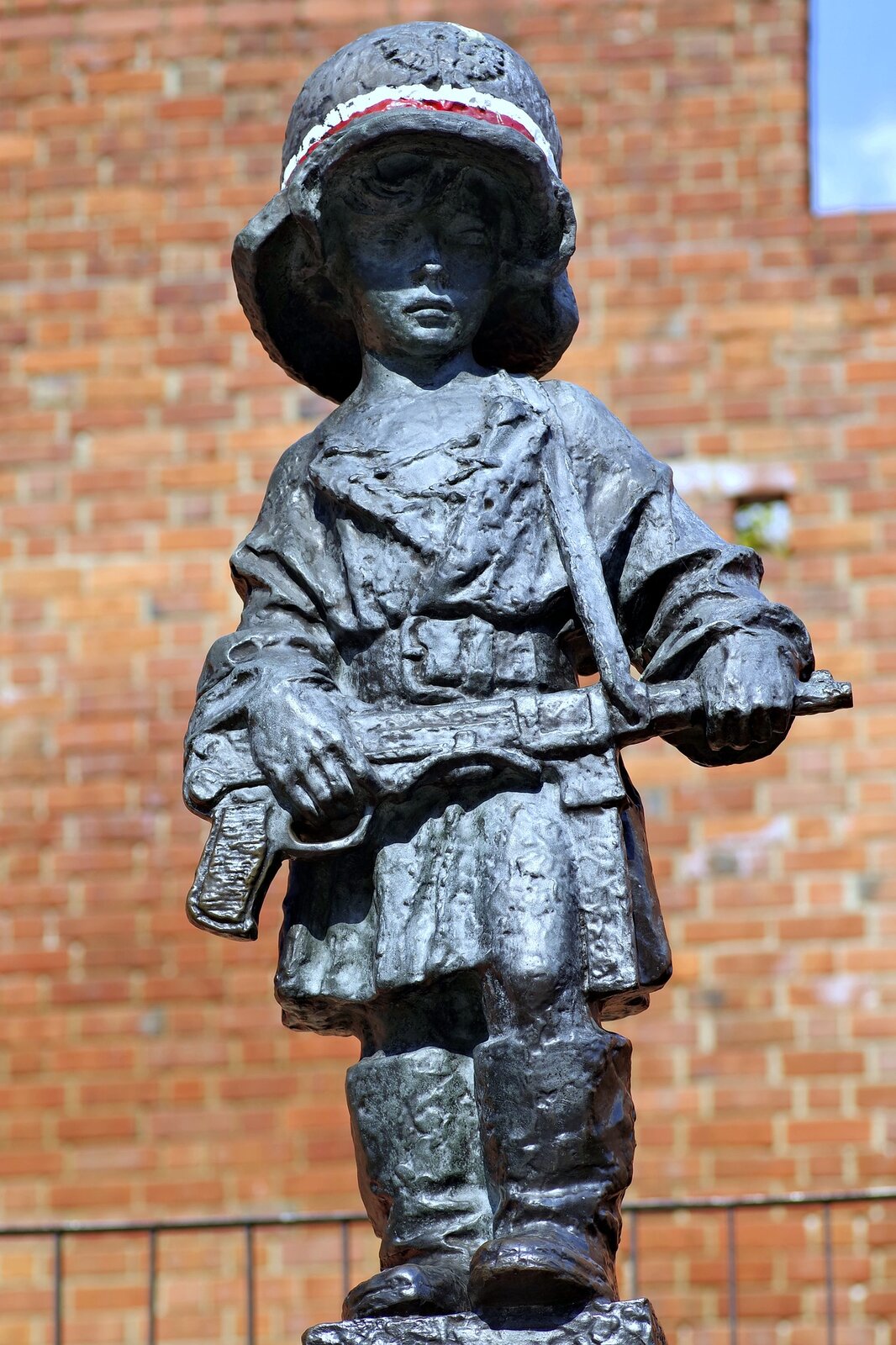 Kolorowa fotografia przedstawia rzeźbę z brązu około 10 letniego chłopca ubranego w za duży płaszcz i buty oraz hełm, na którym widnieje orzeł oraz barwy narodowe Polski. Chłopiec trzyma w dłoniach karabin. Ma poważny wyraz twarzy. W tle ceglany mur. 