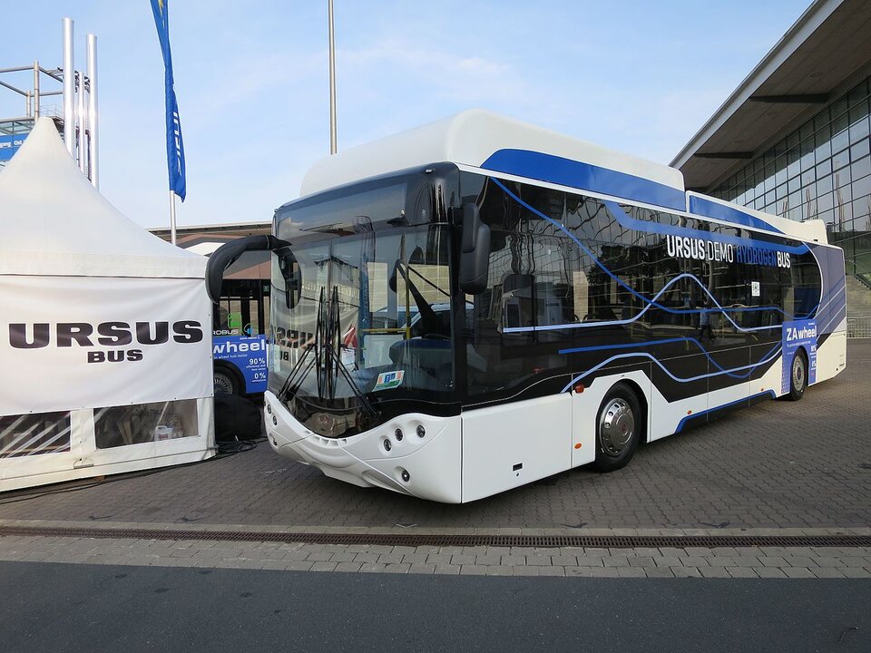 Ilustracja przedstawia  autobus marki Ursus. Jest to nowoczesny autobus zasilany prądem elektrycznym. Jest on w kolorach białym czarnym oraz niebieskim na szybach ma napis Ursus Demo Hydrogen Bus.
