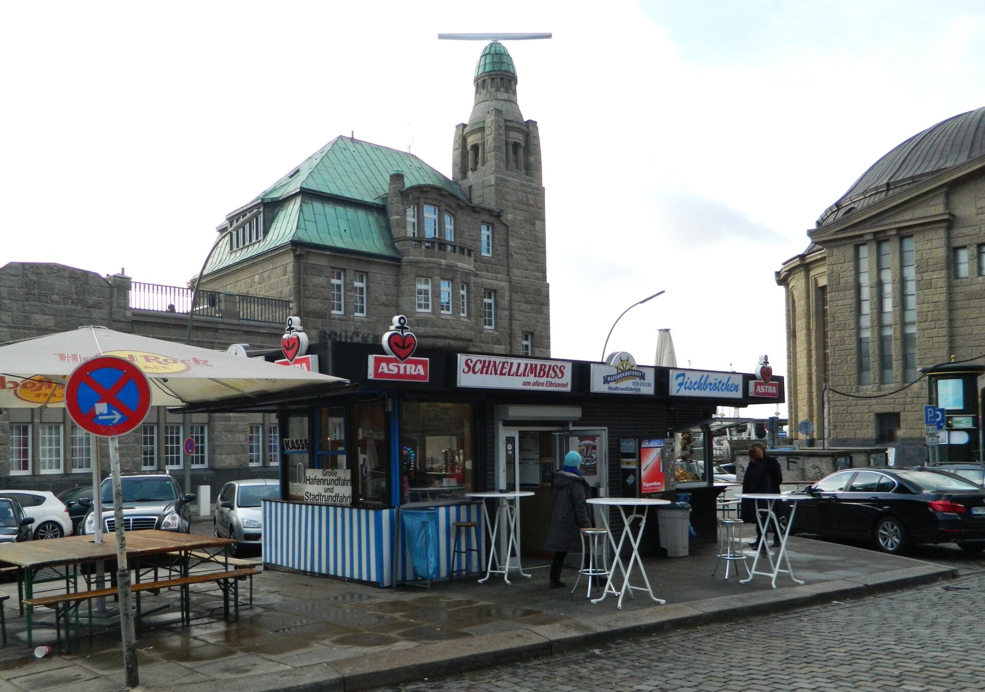 Zdjęcie przedstawia nieduży plac, na którym znajduje się niewielki pawilon z szyldem Schnellimbiss. Przed pawilonem stoją wysokie stoliki oraz ława, a nad nią rozłożony parasol.