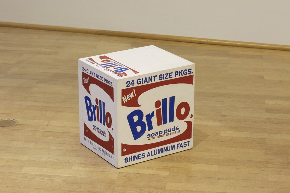 Ilustracja przedstawia fotografię projektu pudełka „Brillo Box”, sporządzonego przez Andy'ego Warhola. Ukazuje ujęcie perspektywiczne pudełka z widoczną ścianą górną i krawędzią boczną. Zawiera napis "Brillo" w niebiesko‑czerwonym kolorze, symetrycznie przedstawiony, ale odwrócony motyw graficzny na dole i u góry w kolorze czerwonym oraz napisy. Wszystkie widoczne ściany boczne są takie same.  