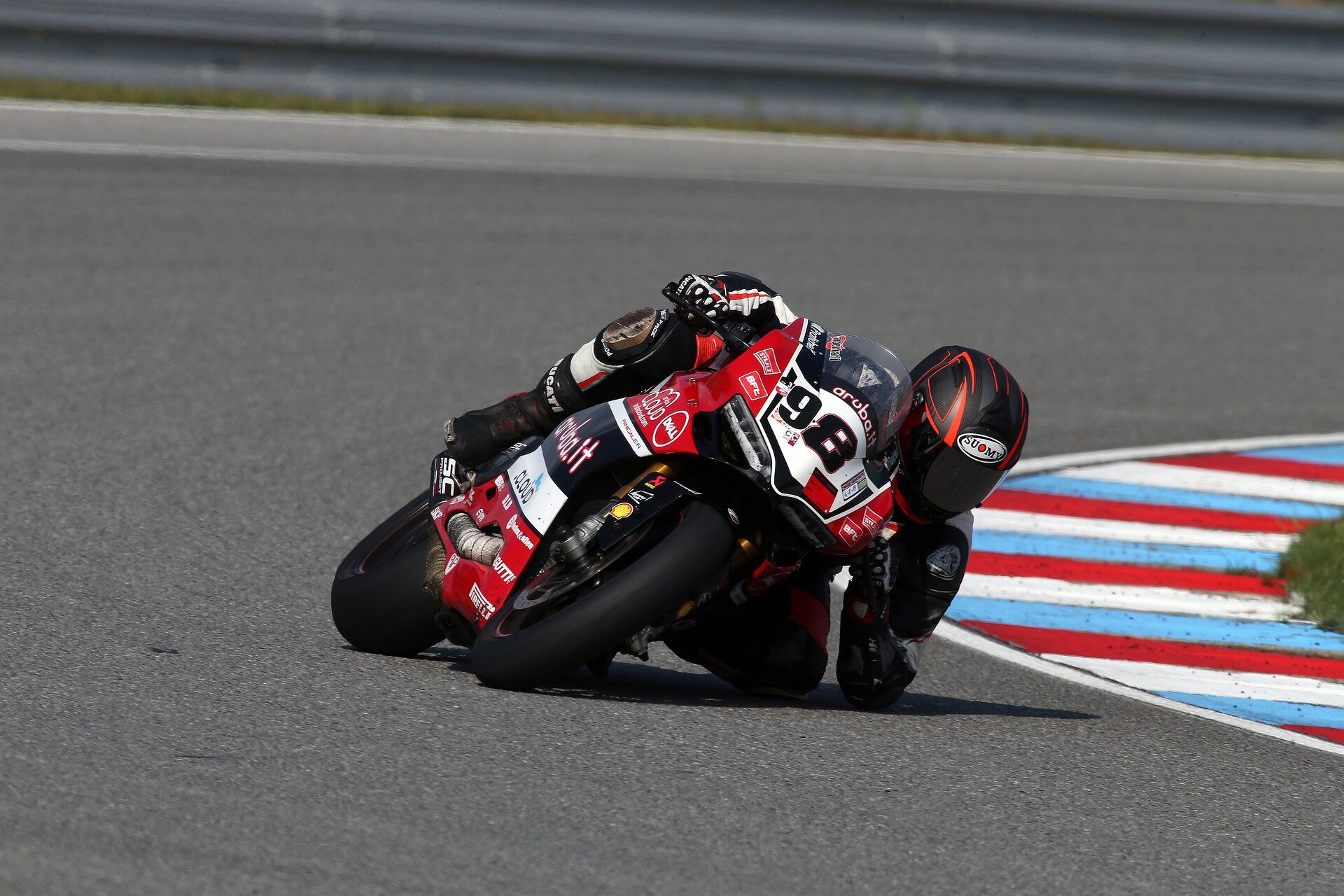 Zdjęcie przedstawia motocyklistę. Jedzie na motocyklu. Jest tak przechylony, że niemal dotyka kolanem i łokciem lewej strony ciała tor, po którym się porusza. Motocykl ma gładkie opony, posiada opływowe osłony.   