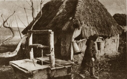 Zdjęcie przedstawia kobietę niosącą wiadro z wodą i cynową kankę na mleko. Wiadro i kana zawieszone są na dwóch przeciwległych końcach kija, który kobieta niesie na ramieniu. Na pierwszym planie widać studnię z drewnianą cembrowiną i kołowrotkiem, służącym do czerpania wody za pomocą wiadra zawieszonego na łańcuchu i spuszczanego w dół. W tle wiejska chata, jednoizbowa, kryta strzechą. Zdjęcie zrobione późną jesienią, zimą bądź wiosną, ponieważ gałęzie drzew pozbawione są liści, a kobieta ubrana jest w walonki na nogach, kufajkę i chustę na głowie. 