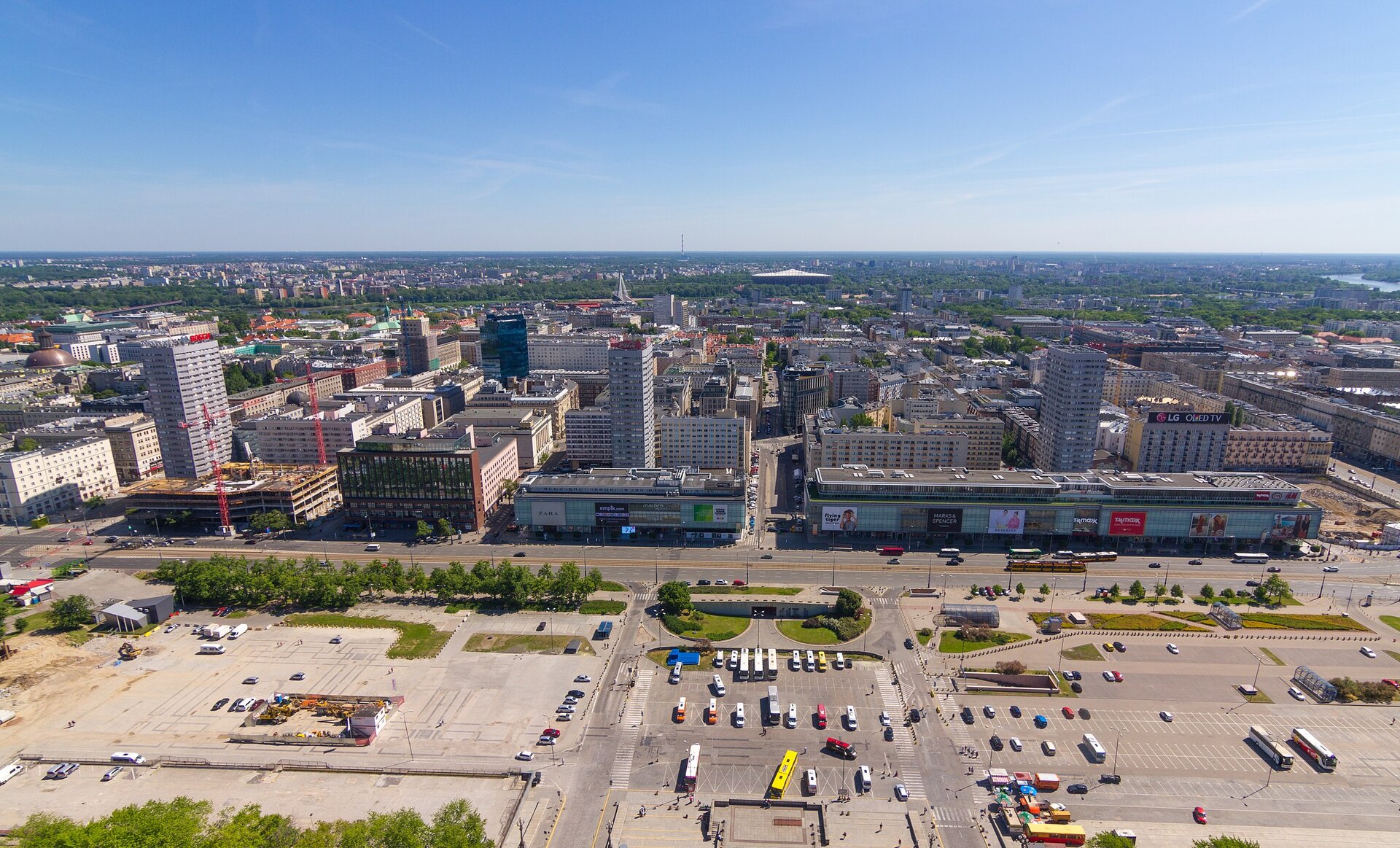 Zdjęcie przedstawia panoramę miasta. Na pierwszym planie znajduje się zabetonowany plac, który w większości wykorzystywany jest jako parking. Za nim ciągną się budynki kamienic oraz blokowisk.