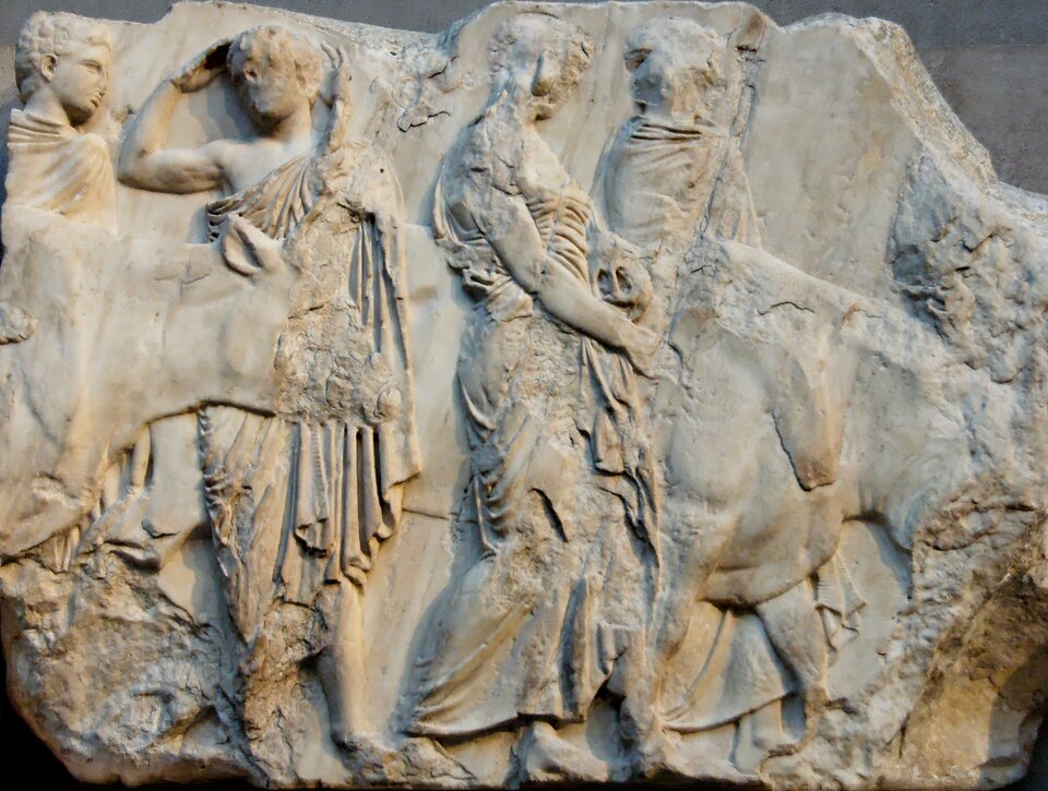 Ilustracja przedstawia fragment Fryzu Panatenajskiego „Woły prowadzone na ofiarę” z południowego fryzu Partenonu, ukazany w dużym zbliżeniu, dzięki czemu wyraźnie widoczne są detale postaci i zwierząt. Po lewej stronie stoi mężczyzna w długiej, ciężkiej draperii, której fałdy układają się w głębokie, pionowe bruzdy; jego sylwetka jest lekko pochylona ku przodowi, jakby kierował ruch procesji. Obok niego widoczny jest drugi mężczyzna, również w długiej szacie, trzymający wodze lub prowadzący konia stojącego tuż przy nim. Koń ukazany jest w ujęciu z profilu, z wyraźnie zaznaczonym łukiem szyi i muskularnym tułowiem; jego głowa jest nieco opuszczona, jakby w spokojnym kroku. Za nim stoi kolejna postać męska, której górna część ciała jest uszkodzona, ale zachowane fragmenty draperii ukazują miękkie, płynne fałdy charakterystyczne dla stylu klasycznego. Po prawej stronie widoczny jest drugi koń, również prowadzony przez mężczyznę; jego sylwetka jest częściowo uszkodzona, lecz zachowane partie ukazują napięcie mięśni i delikatne modelowanie anatomiczne.