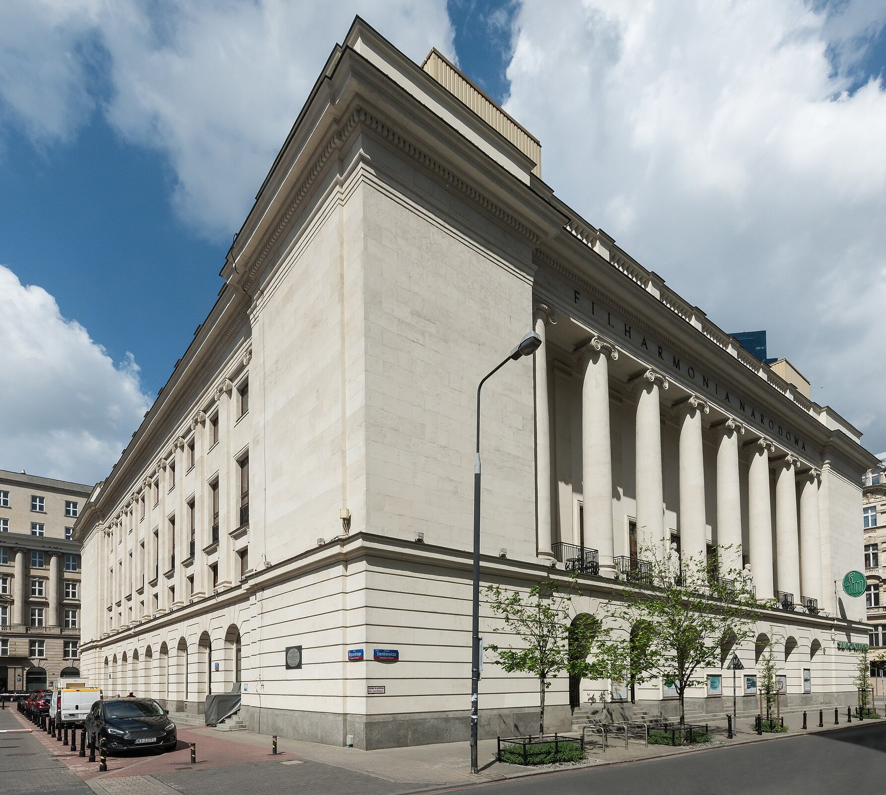 Fotografia przedstawia budynek o biało‑szarej elewacji. Na pierwszym planie znajduje się narożnik budynku, po prawej stronie front Filharmonii. Przód budynku na wysokości pierwszego piętra zdobią masywne kolumny, nad nimi ciemny napis Filharmonia Narodowa. Przed budynkiem rosną małe drzewka.