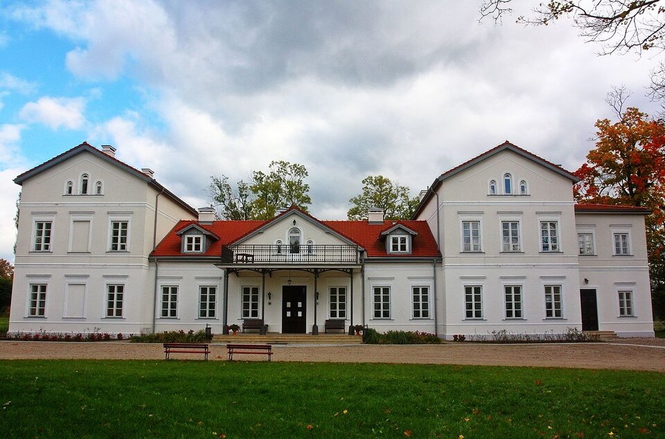 Zdjęcie przedstawia dwór. Na pierwszym planie jest trawnik, potem niewielki dziedziniec ze żwirową drogą. Dalej jest długi budynek dworu. Posiada dokładne wykończenia oraz białą barwę. Dach jest czerwony. Za nim rosną drzewa.