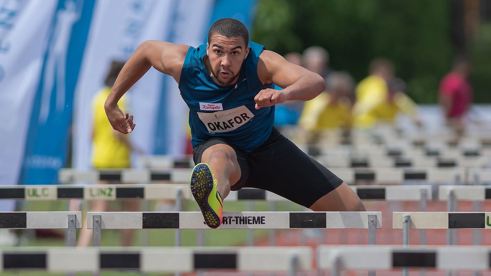 Zdjęcie przedstawia mężczyznę w stroju sportowym, przeskakującego przez płotki lekkoatletyczne. 