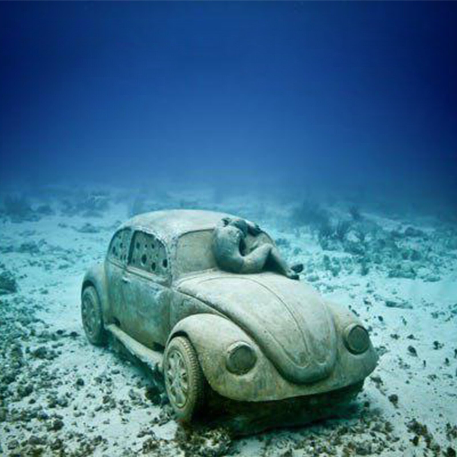 Ilustracja przedstawia rzeźbę samochodu z wybitymi szybami na dnie morza. Na masce samochodu znajduje się oparta o przednią szybę postać, która przypomina dziecko ze względu na swój mały w stosunku do samochodu rozmiar.