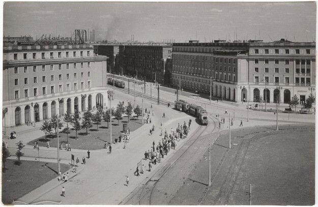 Na zdjęciu znajduje się plac centralny w Nowej Hucie. Widoczne są na nim jeżdżące tramwaje oraz pasy zieleni ze świeżo wsadzonymi drzewami. Przy torach tramwajowych zgromadzeni są ludzie. Na około placu znajdują się nowe bloki i kamienice.