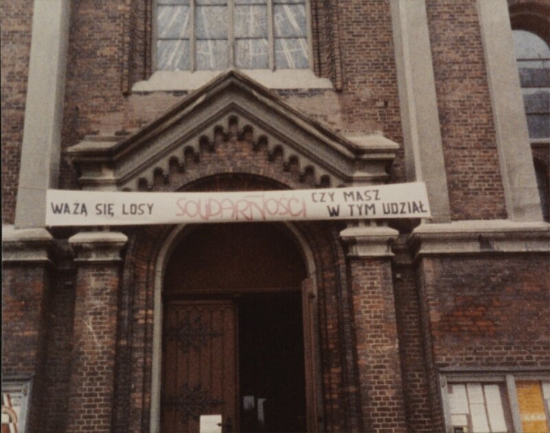 Zdjęcie przedstawia drzwi do kościoła. Nad drzwiami wisi baner z napisem: Ważą się losy Solidarności. Czy masz w tym udział? Kościół jest zbudowany z cegły. 