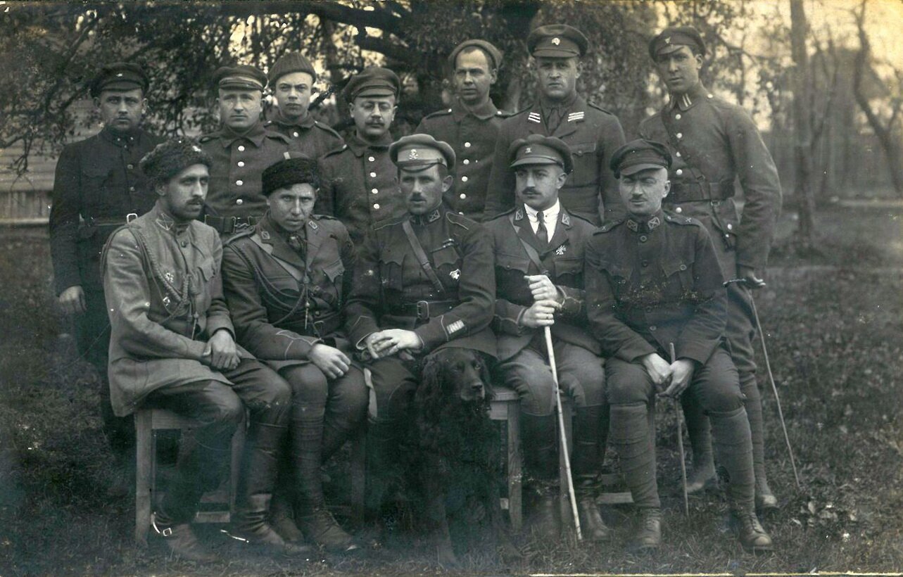 Czarno‑białe zdjęcie przedstawia grupę mężczyzn ubranych w mundury i wojskowe czapki. Pozują oni do zdjęcia na wsi. Część z nich siedzi na drewnianych krzesłach, a reszta stoi za nimi. Niektórzy z nich są uzbrojeni w szable.