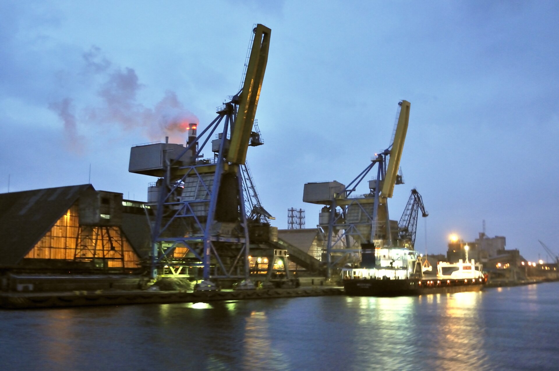 Fotografia kilkudziesięciometrowych żurawi w porcie o zachodzie słońca. Na drugim planie dymiący komin oraz hale fabryczne.