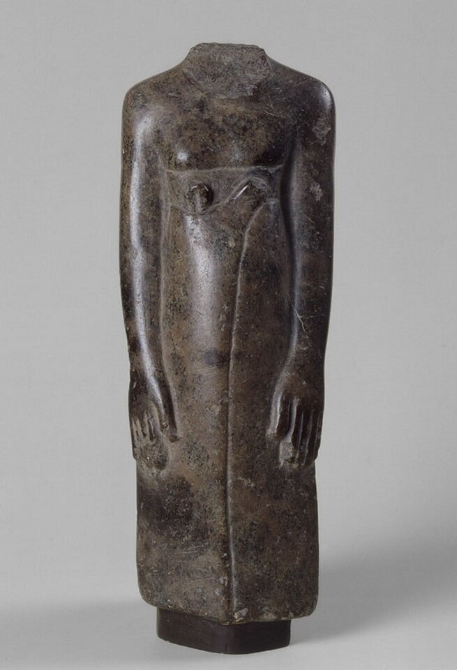 Ilustracjaprzedstawia rzeźbę „Fragment figurki mężczyzny - kapłana”. Widoczna jest stojąca sylwetka postaci bez głowy. Rzeźba wykonana jest prawdopodobnie z diorytu. Ręce są ułożone wzdłuż tułowia. 