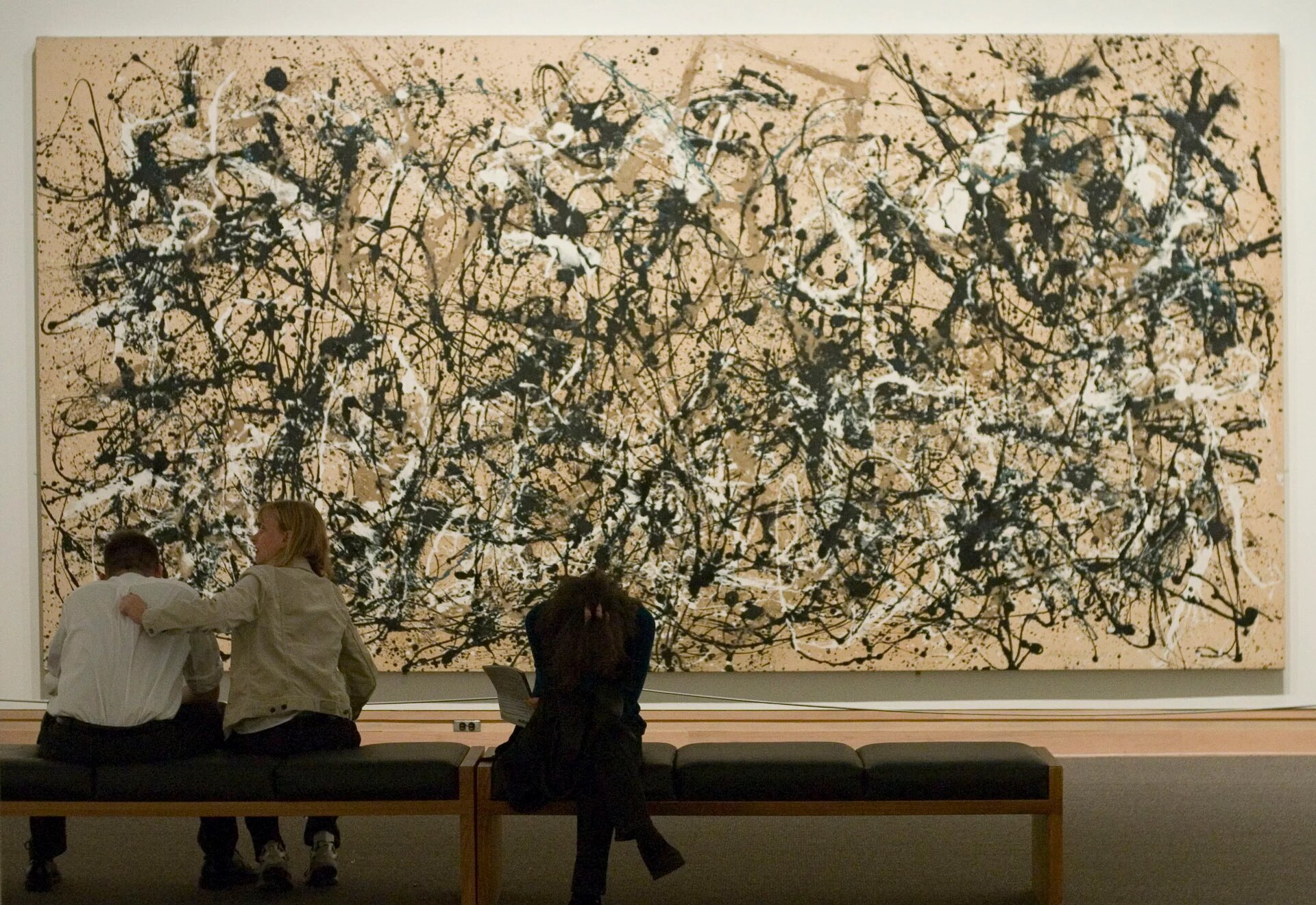 Fotografia przedstawia wielki obraz oraz trzy znajdujące się przed nim osoby. Obraz wykonany jest w stylu abstrakcyjnym, ma układ poziomy i składa się z plątaniny wielu nieregularnych linii różnej grubości, nachodzących na siebie. Obraz jest dynamiczny, bardzo dużo się na nim dzieje, a linie są w kolorze czarnym, białym i brązowym na jasnobrązowym tle.  Pomieszczenie nie jest zbyt jasno oświetlone. Dzieło znajdujące się na ścianie ma układ poziomy i wykonane jest w stylu abstrakcyjnym. Przed obrazem stoją dwie drewniane ławki z ciemnymi, tapicerowanymi siedzeniami. Na ławce znajdującej się po lewej stronie, przodem do obrazu, siedzą dwie osoby - kobieta i mężczyzna. Mężczyzna ma ciemne włosy, ubrany jest w białą bluzę i ciemne spodnie, nogi ma rozstawione szeroko, a łokcie opiera na kolanach. Siedząca obok niego kobieta ubrana jest w beżową kurtkę, granatowe spodnie oraz jasne, sportowe buty. Kobieta trzyma swoją lewą rękę na plecach mężczyzny, a wzrok skierowany ma w lewo. Na drugiej ławce siedzi kobieta skierowana plecami do obrazu. Kobieta ma na sobie ciemne ubrania, nogę założoną na drugą nogę oraz skrzyżowane ręce oparte na kolanach. Głowę pochyla nisko, w jednej ręce trzyma kartkę papieru.