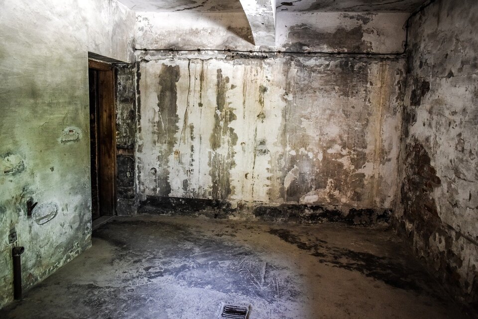 Zdjęcie przedstawia wnętrze pomieszczenia z betonowymi, obdrapanymi ścianami oraz podłogą z metalową kratką po środku. Po lewej stronie są drzwi.