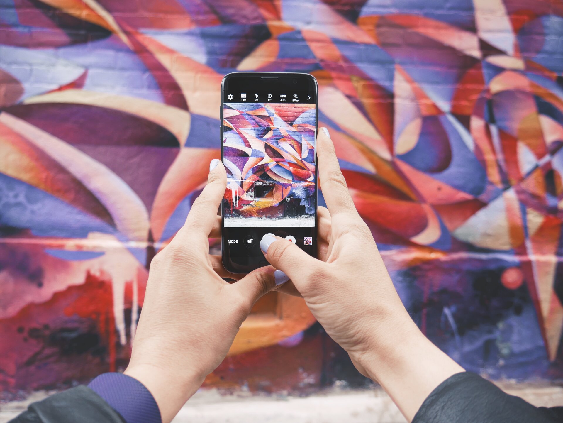 Ilustracja przestawia smartfon w czyichś dłoniach. Ktoś robi zdjęcie pomalowanej na kolorowo ściany znajdującej się w tle.