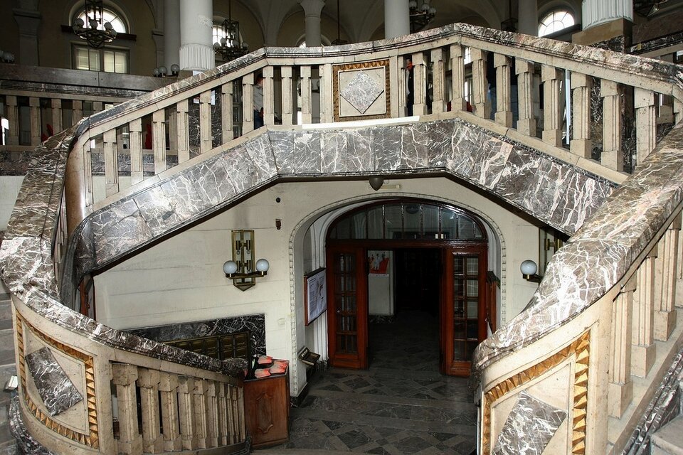 Ilustracja przedstawia Dawny Bank Polski, Gdynia, schody w stylu art déc. Widać przeszklone drzwi, które prowadzą do holu z szaro‑czarną posadzką. z holu prowadzą zaokrąglone marmurowe schody. Wzdłuż schodów ciągną się marmurowe, gęsto ułożone obok siebie kolumny.