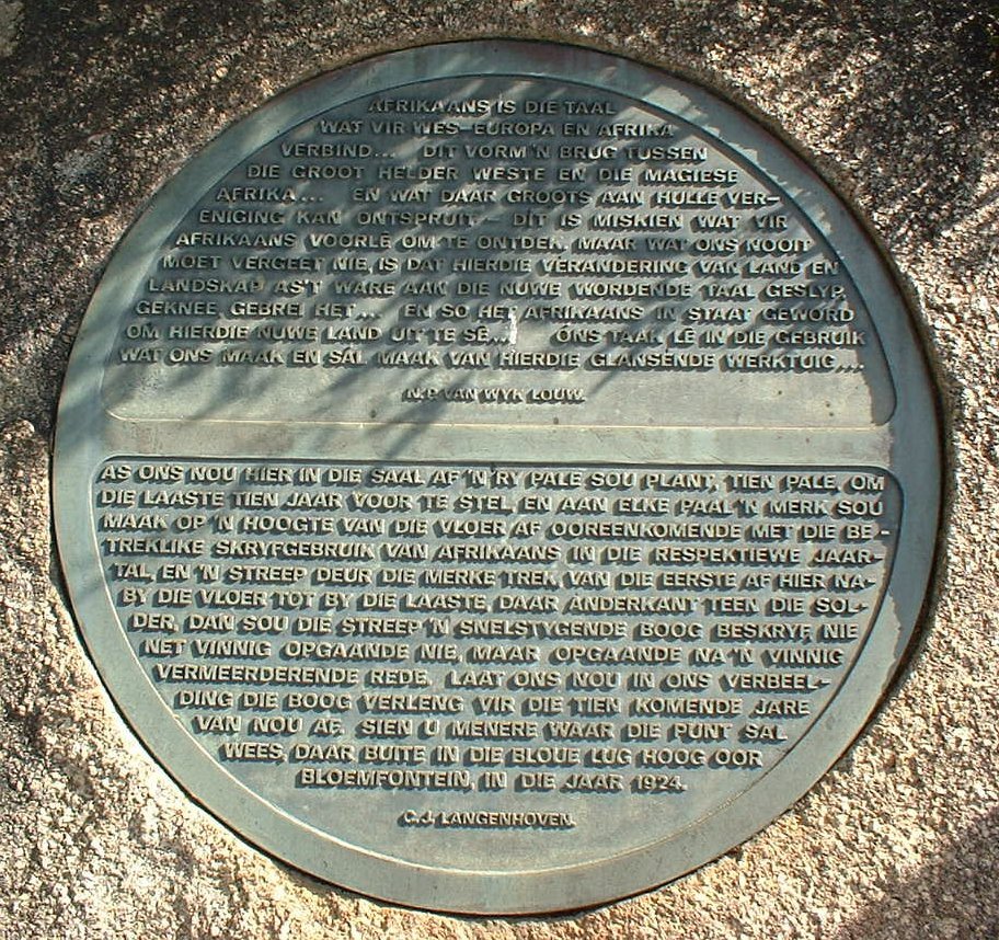 Ilustracja przedstawia płytę z napisami w języku afrikaans używanym przez Afrykanerów. Jest ona wykonana z kamienia, w okrągłym kształcie.