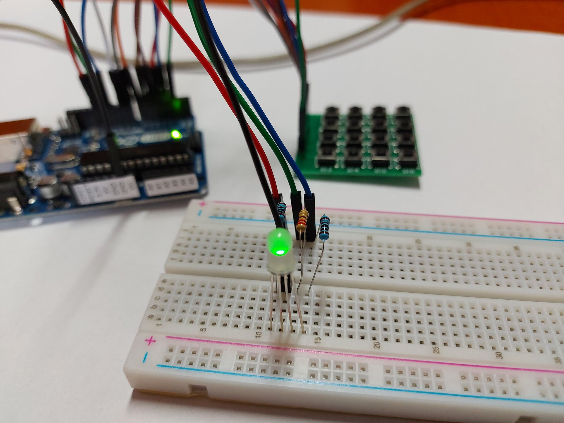 Zdjęcie przedstawia arduino uno połączone kablami do płytki stykowej, na której znajdują się 3 rezystory podłączone do kolejnych nóżek 4 pinowej diody, ostatnia nóżka podłączona jest kablem do arduino. W tle widoczna jest płytka z przyciskami, która podłączona jest do arduino osobnym zestawem kabli. LED świeci się na zielono.