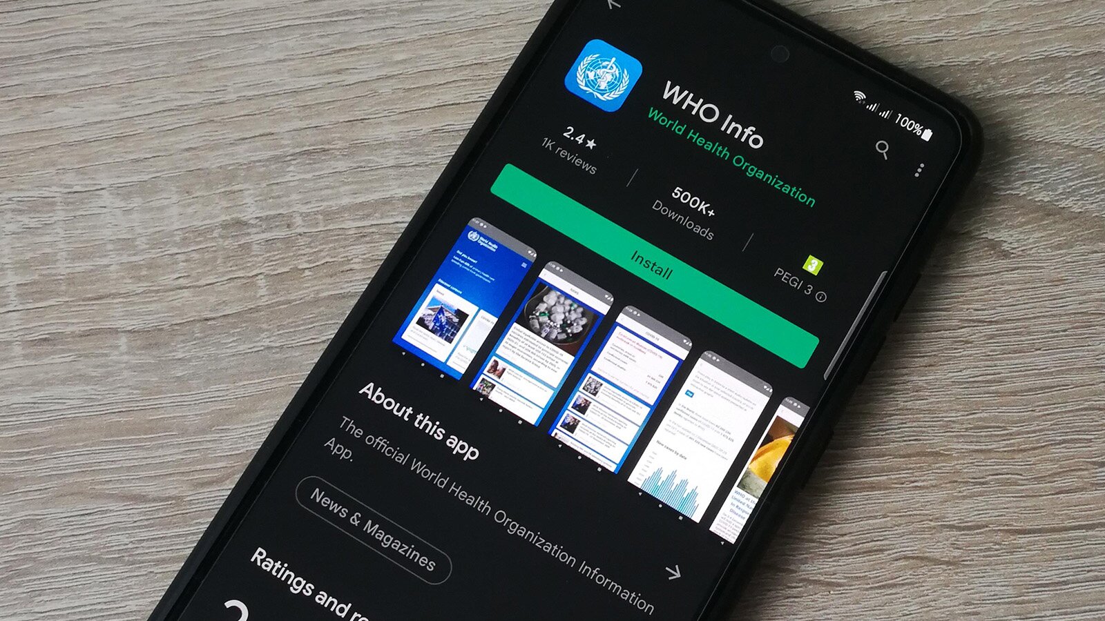 Zdjęcie przedstawia smartfon leżący pod kątem na drewnianym blacie. Na ekranie urządzenia wyświetla się widok aplikacji do pobrania z platformy sklepowej. Widoczne są napisy w języku angielskim, takie jak: WHO INFO, 2.4 1k reviews, 500K Downloads, Install, About this app, The official World Health Organization Information App, News &amp; Magazines.