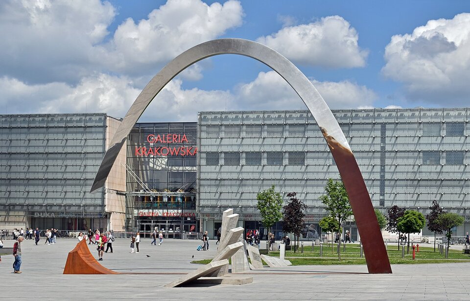 Zdjęcie przedstawia plac przed prostokątnym, kilkukondygnacyjnym, współczesnym budynkiem o szklanej elewacji z napisem nad wejściem: Galeria Krakowska. Na placu stoi pomnik w formie stalowego łuku, który z jednej strony jest postawiony na ziemi, a drugi koniec zawieszony jest nad chodnikiem. Integralną częścią pomnika są podłużne płyty chodnikowe, które są podniesione i wystają poza płaszczyznę placu w formie fali.