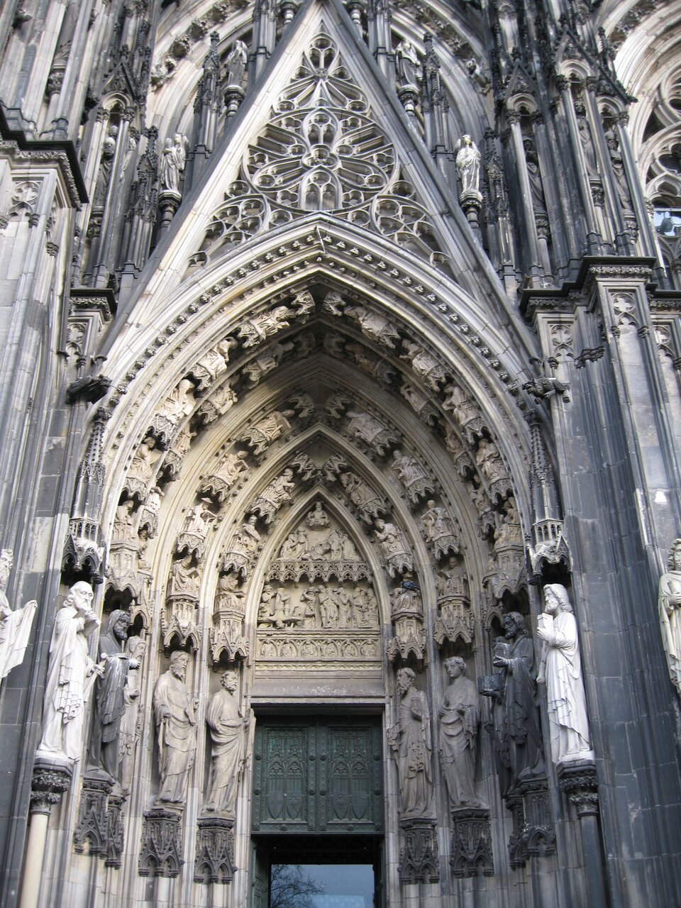 Fotografia przedstawia gotycki portal św. Piotra, fragment fasady zachodniej w Katedrze św. Piotra i Najświętszej Marii Panny w Kolonii w Niemczech. Fasada zachodnia posiada drobne szczegóły architektoniczne oraz rzeźby.