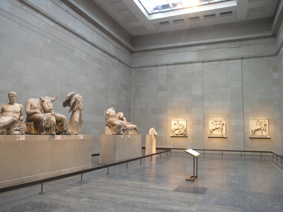 Zdjęcie przedstawia ekspozycję rzeźb z tympanonu Partenonu prezentowaną w Muzeum Brytyjskim w Londynie. W przestronnej, jasnej galerii ustawiono monumentalne figury pochodzące z frontonów świątyni na Akropolu: widoczne są zarówno postacie siedzące i półleżące, jak i fragmenty zwierząt, w tym charakterystyczna głowa konia. Wzdłuż ścian umieszczono także płyty reliefowe z przedstawieniami mitologicznymi. Naturalne światło wpadające przez świetlik podkreśla fakturę marmuru i wydobywa modelunek klasycznych rzeźb, tworząc atmosferę muzealnej prezentacji jednych z najważniejszych dzieł sztuki starożytnej Grecji.