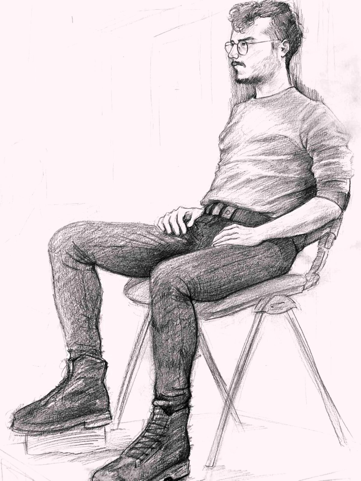 Ilustracja przedstawia realistyczny rysunek młodego mężczyzny siedzącego na krześle. Mężczyzna ustawiony jest do nas bokiem 3/4, ubrany jest w prostą bluzę z podwiniętymi do łokci rękawami i obcisłe spodnie z paskiem. Na nogach ma masywne, sznurowane glany. Mężczyzna siedzi swobodnie, trzymając obie ręce na udach, jedną nogę wspiera się na podwyższeniu. Ma krótkie włosy z kręconą grzywką opadającą na czoło, okrągłe, druciane okulary , niewielkie wąsy i krótką bródkę.