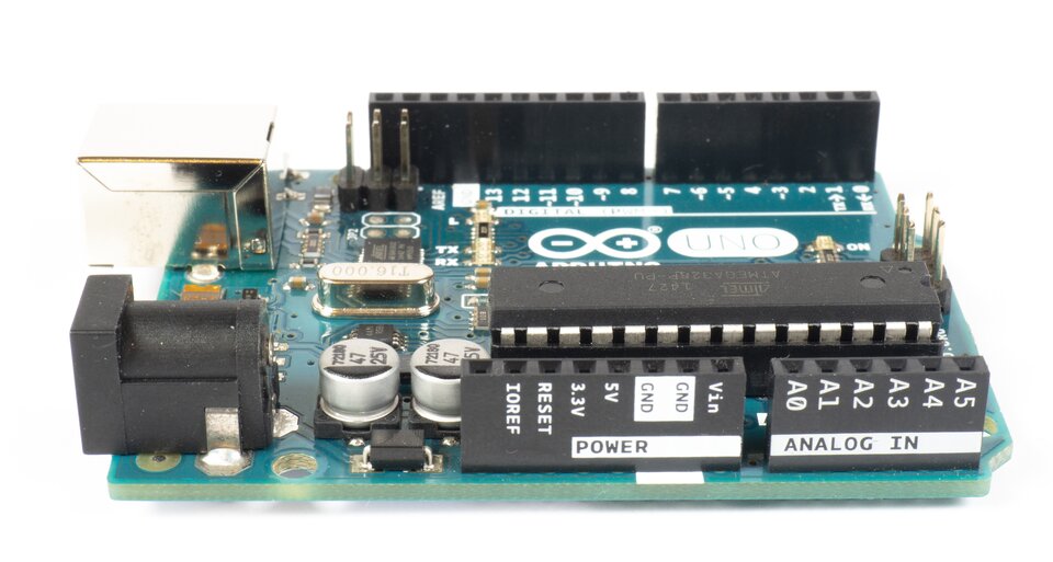 Zdjęcie przedstawia prostokątną, zieloną płytkę, na której zamocowano różne małe elementy. Na płytce są dwa kółka - w środku jednego z nich jest minus, w środku drugiego plus. Obok napis: UNO. Na płytce widać kilka zamocowanych prostokątnych elementów. Na jednym prostokącie jest napis POWER, na drugim ANALOG IN. Prostokąty znajdują się na brzegu płytki, obok siebie.  