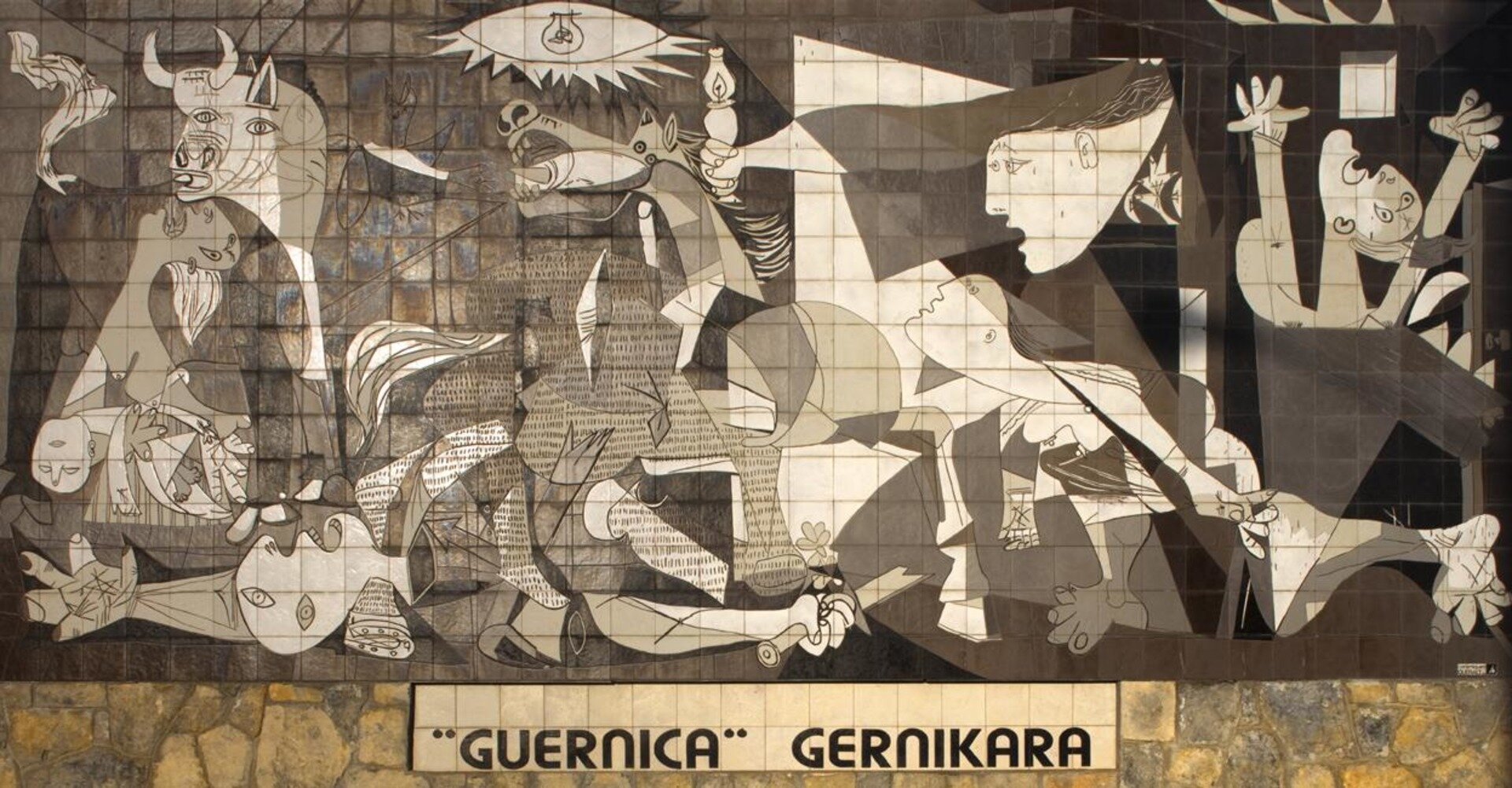 Fotografia barwna przedstawiająca mural "Guernica"-  obraz olejny Pabla Picassa. Obraz przedstawia tragedię hiszpańskiego miasta Guernica, zbombardowanego podczas wojny domowej przez niemieckie lotnictwo. Artysta ukazał chaos, śmierć i strach ludzi, które w spokojnym miasteczku wywołuje wojna. Tragedię Guerniki podkreśla stosowanie przez artystę czerni, bieli i różnych odcieni szarości. Picasso wyraża cierpienie przez postacie i rekwizyty: szczątki ciał, wzniesione do góry ręce i usta otwarte w krzyku, płaczące kobiety, wojownika leżącego ze złamanym mieczem. Symbolicznie obraz ten przedstawia również cierpienie całego narodu baskijskiego walczącego o wolność, o swój kraj i język.