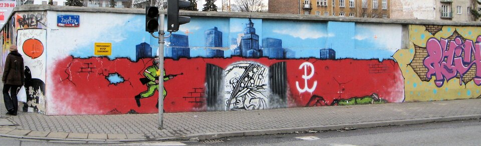 Zdjęcie przedstawia kolorowy mural. Znajduje się na nim scena związana z powstaniem warszawskim. Na tle muru ukazany jest biegnący do bramy powstaniec z karabinem. Na murze widnieje symbol Polski Walczącej.
